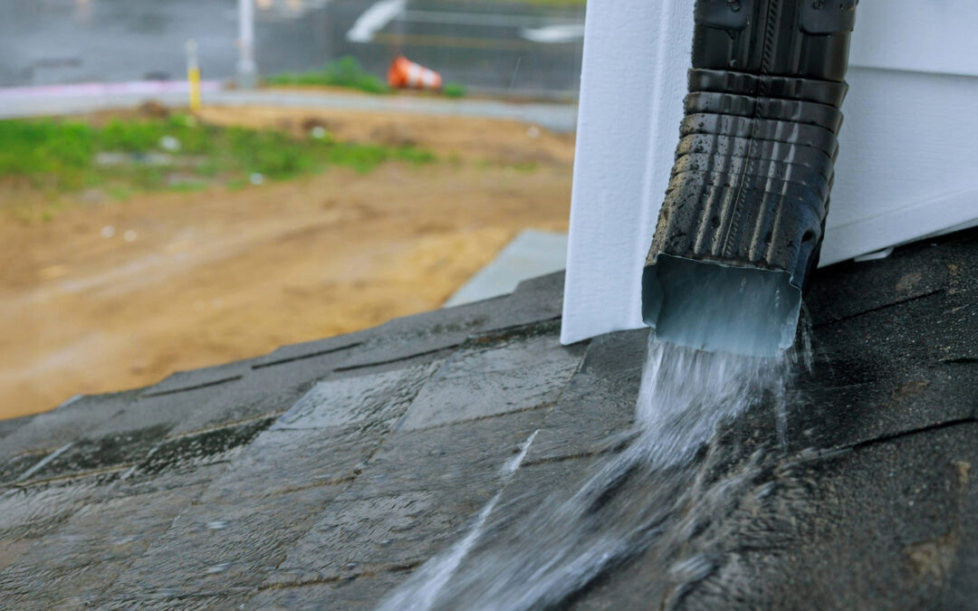 Close-up of rainwater flowing through a black downspout onto a wet asphalt shingle roof during a rainstorm, illustrating effective gutter drainage.