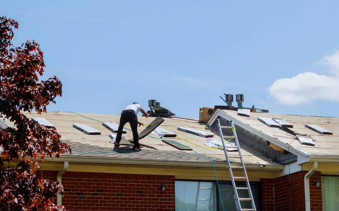 A roofing contractor installing asphalt shingles on a residential home under clear blue skies. The roof is partially stripped with bundles of shingles laid out, and safety ropes are visible. A ladder leans against the house near a brick exterior wall.