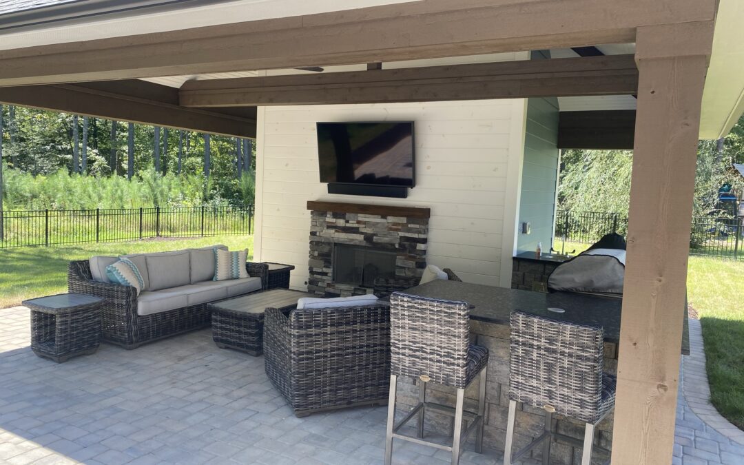 A luxury covered outdoor living space featuring a custom stone fireplace, wall-mounted TV, and a paver patio with wicker seating, showcasing the high-end results homeowners seek when searching for patio companies near me.