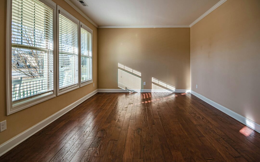 Sunlight streaming through three large windows with white horizontal blinds in an empty room with dark hardwood floors, highlighting the clean finish of a professional window installation.